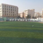 نمای کلی زمین ورزشی سبز محصور با درختان و ساختمان‌های اطراف در پس‌زمینه