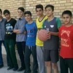 گروهی از دانش‌آموزان ایستاده کنار دیوار آجری در حیاط مدرسه با یک معلم
