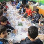 دانش‌آموزان در حیاط مدرسه در حین انجام یک فعالیت رنگ‌آمیزی و کار هنری جماعی بر روی یک سطح بزرگ