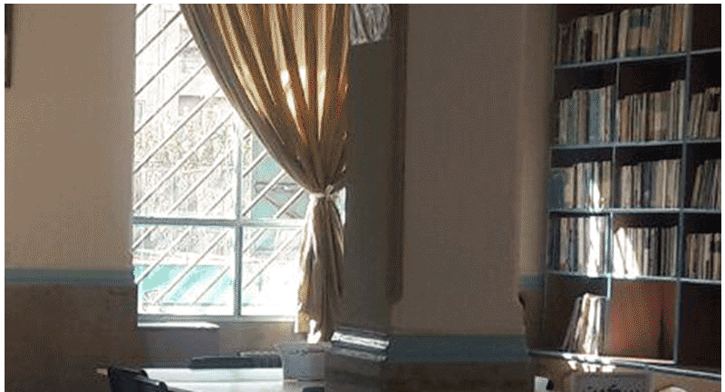 فضای کتابخانه مدرسه با قفسه‌های کتاب، میز کار چوبی، پنجره‌ای بزرگ با نور طبیعی و پرده قهوه‌ای