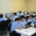 دانش‌آموزان پسر با لباس مدرسه در حال کار روی کامپیوترها (لپ‌تاپ و دسک‌تاپ) در سایت کامپیوتر مدرسه