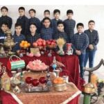 دانش‌آموزان و معلمی در مراسم جایزه‌دهی سالانه کنار میزی تزیین‌شده با کاپ‌های طلایی و برنجی، گل‌های قرمز و تزیینات تحصیلی