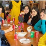 گروهی از دانش‌آموزان در یک رویداد اجتماعی دور میز غذایی، میز با تزئینات گلی و مشروبات شاد و صمیمی