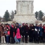 گروهی از دانش‌آموزان و معلمان در یک مراسم دسته‌جمعی پشت یادمان عمومی، فضای باز