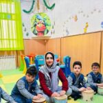 معلم و دانش‌آموزان در کلاس درس، نشسته در حال فعالیت آموزشی با سازهای موسیقی و تزئینات دیواری تعلیمی