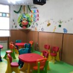 کلاس درس کودکان مجهز به میزهای رنگی و صندلی‌های متناسب، دیوارهای تزیین‌شده با نقاشی‌های بزرگ و اشکال هندسی رنگین