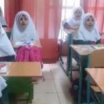 کلاس درسی با دانش‌آموزان دختری در حال نشستن روی صندلی‌ها و میز‌های درسی در اطاق درس