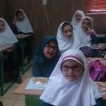 کلاس درسی با دانش‌آموزان دختر نشسته در میزهای فردی، حضور معلم و تخته‌سیاه در پس‌زمینه