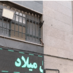 نمای بیرونی ساختمان مدرسه با درهای آهنی و پنجره‌های حفاظت‌شده و تابلوی نیون سبز نام مدرسه میلاد