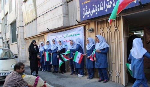دانش‌آموزان با لباس مدرسه‌ای و پرچم‌های رنگی ایران در جلوی درب مدرسه در یک جشن/مراسم ملی