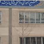 نمای خارجی ساختمان مدرسه با تابلوی دائمی فارسی در بالا، پنجره‌های فلزی‌شده و درخت در کنار ساختمان