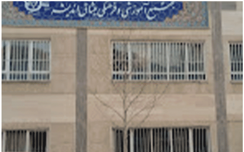 نمای خارجی ساختمان مدرسه با تابلوی دائمی فارسی در بالا، پنجره‌های فلزی‌شده و درخت در کنار ساختمان