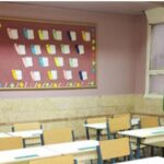 نمای داخلی کلاس درس با میز‌های چوبی دانش‌آموزی، صندلی‌های تک‌نفره، تخته دیواری تزیینی با نقاشی‌های رنگی و پرده سبز