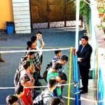 دانش‌آموزان و معلمان در حیاط مدرسه در حال شرکت در فعالیتی گروهی، به احاطه دیوارها و درب‌های رنگی