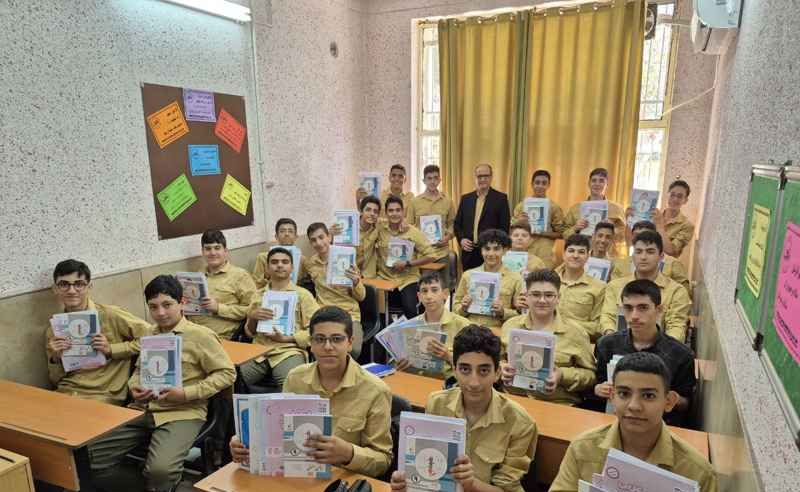 دانش‌آموزان پسر با یونیفرم زرد در حال دریافت کتاب‌های علمی از دست معلم در کلاس درس