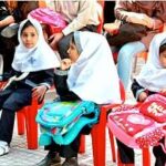دانش‌آموزان در حیاط مدرسه نشسته اند و کیف‌های رنگین کوله‌پشتی را در مراسم رسمی توزیع دریافت می‌کنند