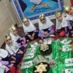 گروهی از دانش‌آموزان با تاج‌های کاغذی رنگین در حال جشن‌گرفتن و خوردن میان‌وعده‌های متنوع روی فرش سبز
