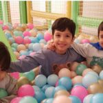 سه دانش‌آموز در حال بازی و سرگرمی در سالن بازی داخلی پر از توپ‌های بادی رنگی با لبخند‌های شادی