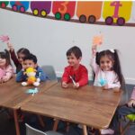 گروهی از دانش‌آموزان کوچک در یک میز چوبی نشسته‌اند و هر کدام در حال ساخت و کار دستی با مواد رنگی هستند، دیوار پشت آن‌ها تزئین شده است با رنگ‌های شاد و نقاشی‌های هندسی.