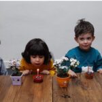 چهار دانش‌آموز جوان در حال انجام فعالیت عملی کاشت و پرورش گل‌های کوچک در گلدان‌های رنگی، دور یک میز چوبی