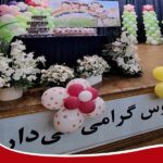 صحنه تزئینی برای یک مراسم یا جشن مدرسه‌ای با بالون‌های صورتی و سبز، گلهای سفید، و متن فارسی روی پارچه‌نوشته