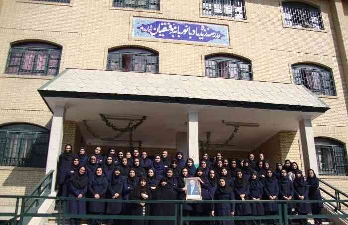 گروهی از دانش‌آموزان دختر مدرسه در سایه‌روی ساختمان برای یک مراسم یا رویداد خاص جمع شده‌اند، با تابلوی فارسی مدرسه به شکل چشم‌گیری مرئی