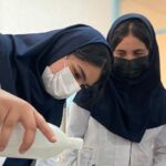 دو دانش‌آموز دختر با لباس آزمایشگاه و ماسک در حین انجام آزمایش علمی