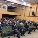 گردهمایی دانش‌آموزان و بزرگسالان در سالن مراسم با صندلی‌های سبز و صفحات نمایش دیجیتال