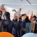دانش‌آموزان دبستانی در حال رای‌دهی در مراسمی داخل کلاس درس با بالون‌های تزیینی نارنجی و کاغذ‌های رنگی