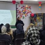 دانش‌آموزانی نشسته در کلاس‌درسی در حال حضور در یک مراسم یا سخنرانی با تزئینات رنگی و صفحه‌نمایش پس‌زمینه