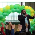 مراسمی در مدرسه با یک سخنران مرد در حال سخنرانی کنار بالون‌های سبز و زرد و دانش‌آموزانی با دستان بالا