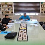چهار نفر در حال بحث و کار بر روی اسناد و کاغذهای رنگی اطراف میز سفید در سالن با تابلو و قفسه‌های چوبی