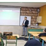 معلمی در حال سخنرانی به دانش‌آموزان در حالی که آنها در میز‌های سبز رنگ نشسته‌اند و به سمت تخته و پرده پیش جلو گوش می‌دهند