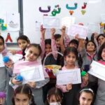گروهی از دانش‌آموزان در کلاس درس که کارهای هنری و کاردستی‌های رنگی را نشان می‌دهند و همدیگر را جشن می‌گیرند، فضایی پر از میز، تزیینات دیواری و تصاویر هنری.