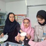 دانش‌آموزان دختر در حال انجام یک آزمایش علمی عملی با نظارت معلم در آزمایشگاه مدرسه. یکی از شاگردان یک ظرف شفاف سفید در دست گرفته است.