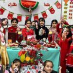 گروهی از دانش‌آموزان پسر و دختر با لباس‌های رنگی و سنتی، در حال شرکت در مراسم شب یلدا یا جشن‌گیری درون‌مدرسه‌ای، با تزیینات و نوار‌های قرمز روی دیوار و میز تزیین‌شده