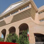 نمای خارجی سردر ساختمان مدرسه با معماری مدرن آجری، پنجره‌های افقی صفحه‌ای و درب ورودی مرتفع، تزئینات معماری کلاسیک در بالای ساختمان، و پرچم ملی ایران.