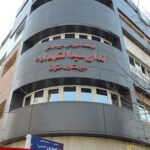 نمای خارجی ساختمان مدرسه با تابلوی نام فارسی، ورودی با درب و پنجره‌های بزرگ و تابلوی آبی مطالبات.