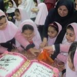 دانش‌آموزان دختر با حجاب صورتی و سیاه در حال جشن‌گیری اطراف کیک‌های تزیین‌شده با نوشته‌های فارسی در مراسم مدرسه