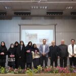 گروهی از دانش‌آموزان و معلمان در حین یک مراسم رسمی پشت گل‌های تزیینی و پرده نمایش داخل تالار