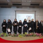 گروهی از دانش‌آموزان ایستاده در حال شرکت در یک مراسم رسمی در سالن مدرسه، با صفحه نمایشی در پس‌زمینه