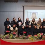 گروهی از دانش‌آموزان دختر حجاب‌پوش در حال حضور در یک مراسم یا رویداد مدرسه‌ای با تزیینات گل‌های قرمز و صفحه نمایش در پس‌زمینه
