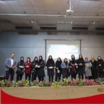 مراسم رسمی اختتام با حضور معلمان و دانش‌آموزان ایستاده روی صحنه، تزئین شده با گل‌های قرمز