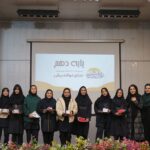 گروهی از دختران دانش‌آموز با لباس رسمی و حجاب در حال شرکت در مراسم رسمی درون‌سالن، پشت‌شان پرده نمایشی با لوگو و تزئینات گلی