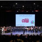 دانش‌آموزان روی صحنه در مراسمی جشن‌واره برای دریافت نشان‌های تقدیری در رویداد آموزشی و پرورشی شنایی، با صحنه‌ی روشن‌شده و حضور مهمانان در سالن بزرگ