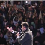 یک مدرس یا سخنران جلوی جمع تعداد زیادی دانش‌آموز صحبت می‌کند و دست‌هایش را بالا می‌گیرد