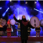دانش‌آموزان با لباس‌های سنتی رنگارنگ در حال اجرای موسیقی به‌کمک ساز‌های دستی تقلیدی روی صحنه با نورپردازی آبی متحرک