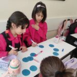 دانش‌آموزان دختر در کلاس درس، در حال انجام یک فعالیت علمی تجربی با رسم دایره‌های آبی روی کاغذ و استفاده از مارکرها