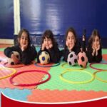 چهار دانش‌آموز در حال بازی و تعامل با توپ‌های فوتبال روی زمین رنگی سالن ورزشی