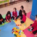 دانش‌آموزان در حیاط مدرسه حول اشکال رنگین و لگو نشسته‌اند و روی پروژه‌ی ساختمانی/مهندسی با یکدیگر کار می‌کنند.